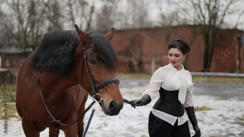 strict horsewoman is leading brown steed in horse yard in winter day