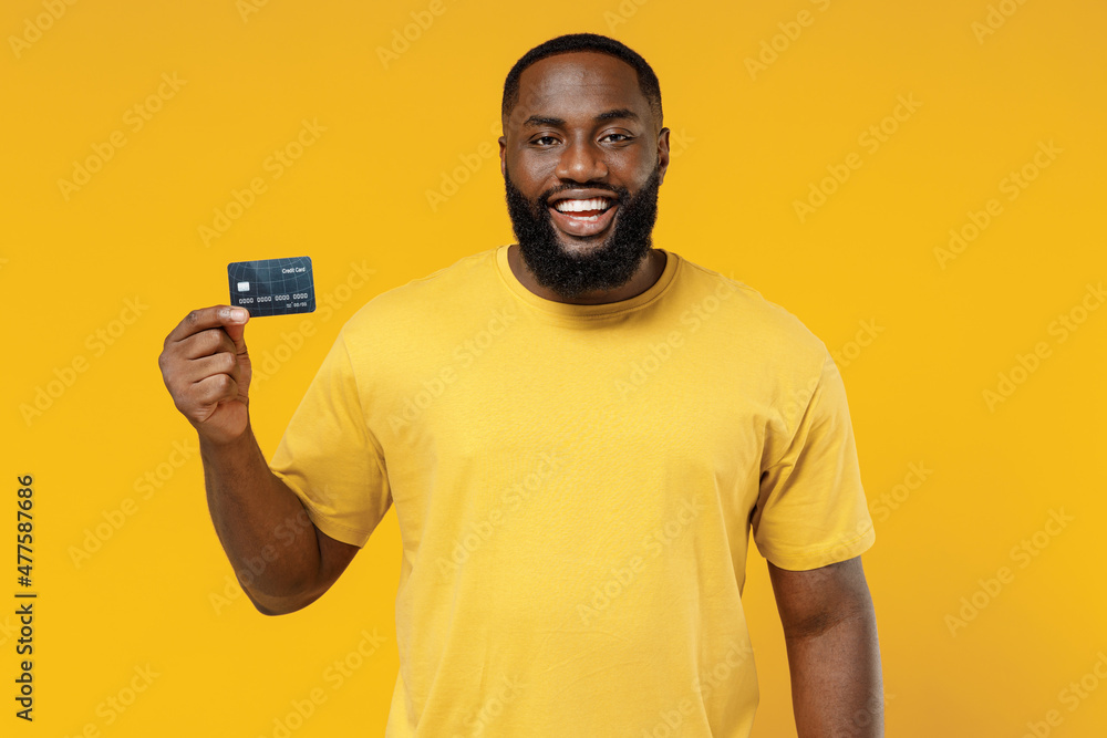 Young smiling satisfied fun rich cool happy black man 20s wear bright casual t-shirt hold in hand credit bank card isolated on plain yellow color background studio portrait People lifestyle concept