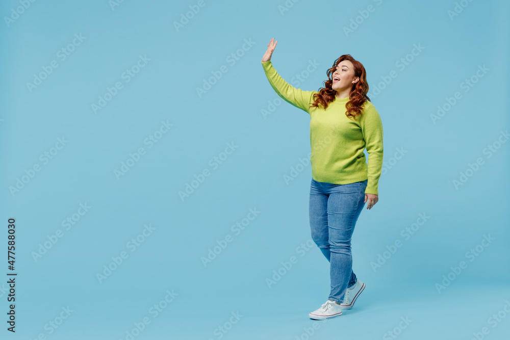 Full body friendly happy young chubby overweight plus size big fat fit woman wear green sweater walking going waving hand isolated on plain blue background studio portrait. People lifestyle concept. Full body friendly happy young chubby overweight plus size big fat fit woman wear green sweater walking going waving hand isolated on plain blue background studio portrait. People lifestyle concept.