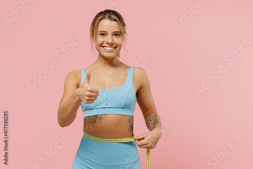 Young fun sporty athletic fitness trainer instructor woman wear blue tracksuit spend time in home gym hold measure tape on waist show thumb up isolated on plain pink background. Workout sport concept.