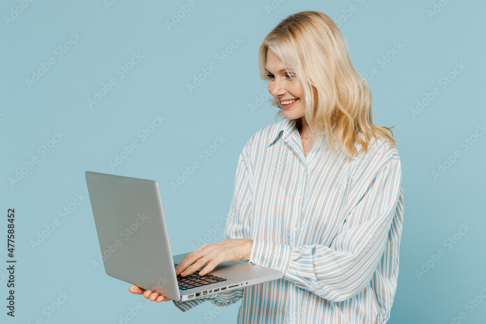 Elderly smiling happy fun caucasian woman 50s wearing striped shirt hold use work on laptop pc computer isolated on plain pastel light blue color background studio portrait. People lifestyle concept. Elderly smiling happy fun caucasian woman 50s wearing striped shirt hold use work on laptop pc computer isolated on plain pastel light blue color background studio portrait. People lifestyle concept.