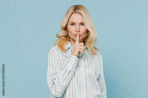 Elderly secret caucasian woman 50s wear striped shirt say hush be quiet with finger on lips shhh gesture isolated on plain pastel light blue color background studio portrait. People lifestyle concept.