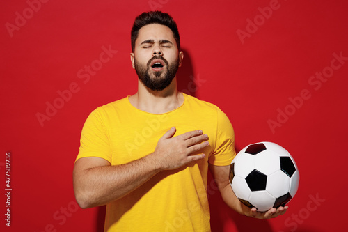Foto Vivid young bearded man football fan in yellow t-shirt cheer up support favorite team eyes closed hold soccer ball sing national country anthem isolated on plain dark red background studio portrait