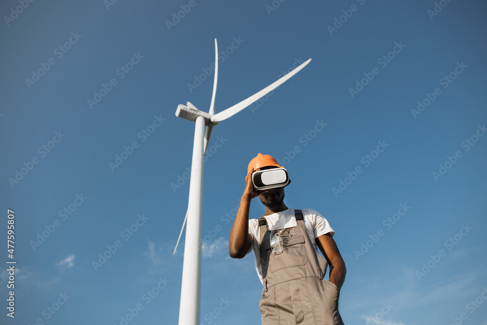 African american man wearing VR headset while standing at rural area with huge windmills around. Low angle view. Technology and people concept. African american man wearing VR headset while standing at rural area with huge windmills around. Low angle view. Technology and people concept.