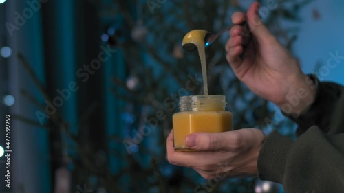 holding a jar of honey against the background of a Christmas tree. wrap thick honey on a spoon in slow motion.