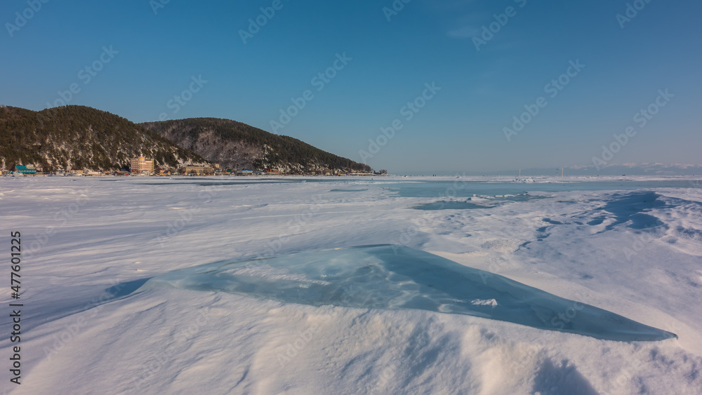 Ice and snow lie on the surface of a frozen lake. City houses are visible on the shore. A wooded mountain range against a blue sky. Baikal