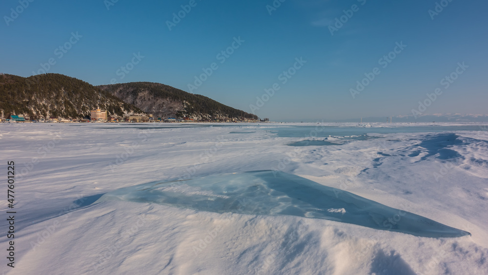 Ice and snow lie on the surface of a frozen lake. City houses are visible on the shore. A wooded mountain range against a blue sky. Baikal Ice and snow lie on the surface of a frozen lake. City houses are visible on the shore. A wooded mountain range against a blue sky. Baikal