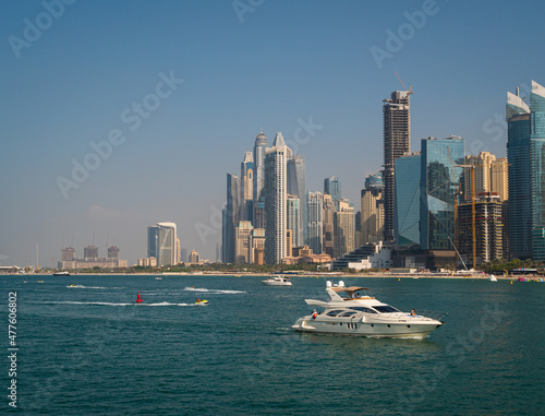 Leisure sailing boats cruise blue waters along the sandy shoreline of Jumeirah Beach at Dubai with high rise luxury hotels and waterfront apartment towers in the background beneath a clear blue sky.