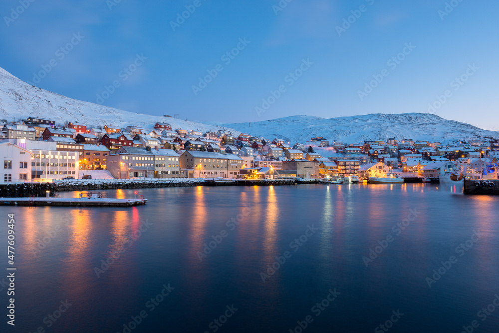 A horizontal landscape of a Norwegian coastal town, with lights reflecting of the water, taken from a boat on the sea in the early evening, Norway