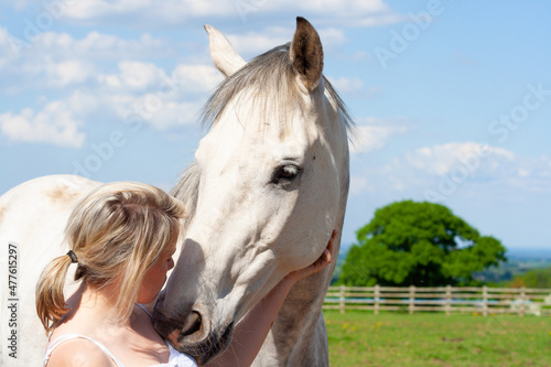 Pretty young woman and her beautiful dapple grey horse share a loving moment outdoors in field in English countryside on a summers day.