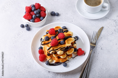 Viennese or Belgian waffles with fresh berries (raspberries and blueberries) and chocolate sauce on a white plate and a cup of coffee. Traditional dessert. Close-up, white background.