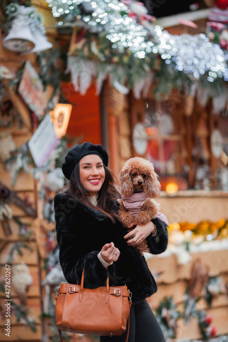 Happy emotional young dark-haired woman in faux fur coat and beret shopping with her dog apricot little poodle on the street of a European city. Winter holidays. Christmas.