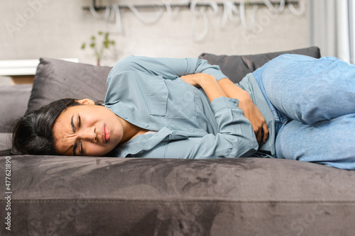 Sad young multiracial woman wearing casual clothes suffering from menstrual pain, feeling sick to her stomach, holding belly, having abdominal cramps during period and lying down on bed at home © Vadim Pastuh