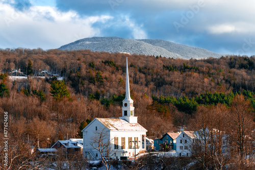 Small village surrounded by pine and maple tree forest with snow covered mountains in background.