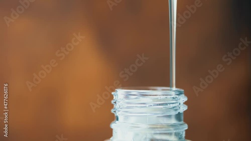 Thick transparent liquid slowly pours in thin stream. Plastic bottle neck. Syrup spreads along thread. Liquid soap or gel dish drips into container. Close-up side. Unknown chemical substance is poured