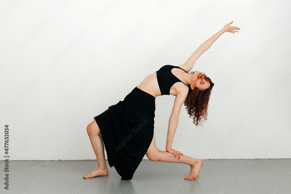 Young redhead woman dancer performing dance move, leaning on her led, stretching her arm up, skirt posing on a white studio background