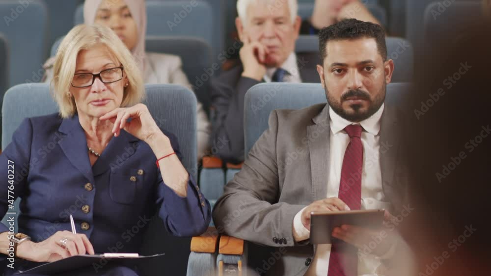 PAN shot of diverse male and female politicians listening to speaker at political forum in big conference hall PAN shot of diverse male and female politicians listening to speaker at political forum in big conference hall