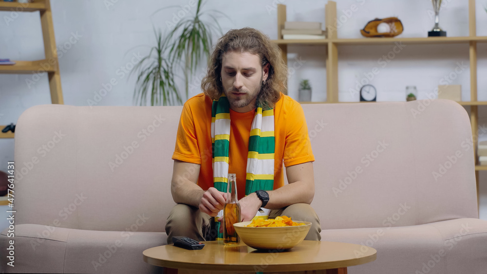 sad sport fan in striped scarf sitting on couch near tv remote controller, beer and chips on coffee table. sad sport fan in striped scarf sitting on couch near tv remote controller, beer and chips on coffee table.