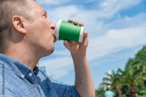 Man drinks coffee from a green paper cup with a place for text against the sky and palm trees, copy space