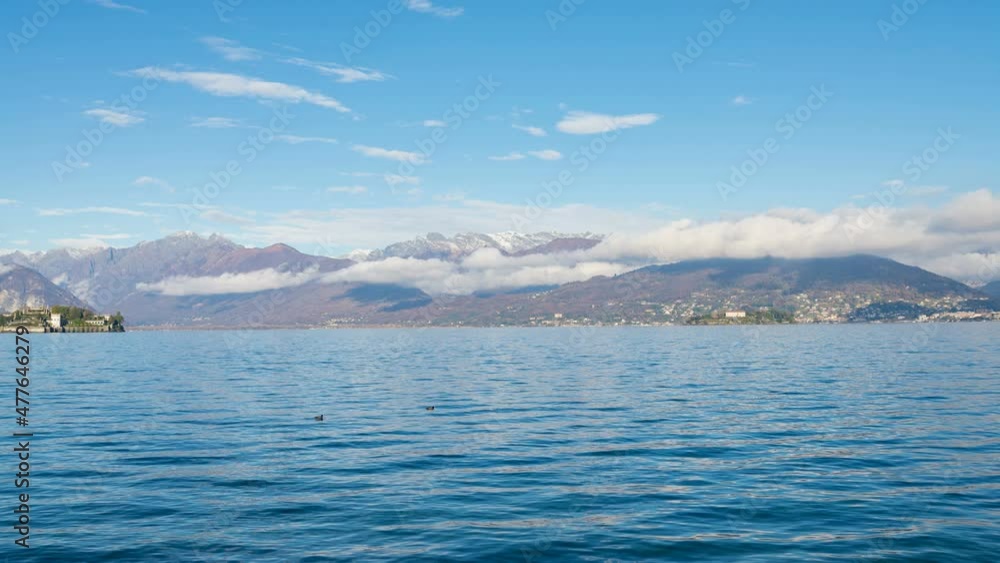 The blue waters of Lake Maggiore, from a small village on the coastline named Stresa. Italian alps on the background, with moving white clouds and blue sky. Time lapse. The blue waters of Lake Maggiore, from a small village on the coastline named Stresa. Italian alps on the background, with moving white clouds and blue sky. Time lapse.
