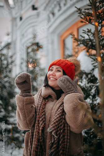 Portrait of a young smiling brunette girl in an orange hat and a beige fur coat on the street against the background of Christmas trees with a lit sparkler