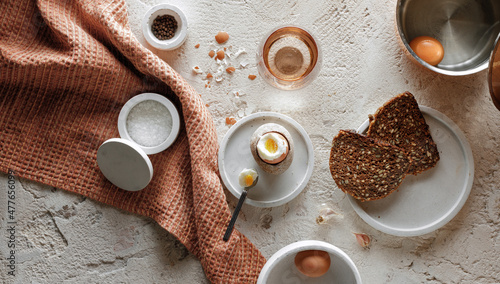 Breakfast setting with boiled egg in stone egg cup, whole grain rye bread with seeds, glass of water, salt flakes and pepper in concrete bowls on textured clay background. Healthy breakfast concept.