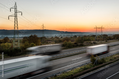 Fototapeta Naklejka Na Ścianę i Meble -  High spped blurred highway traffic with Delivery Lorry trucks passing in both directions. Intense Highway transportation with delivery trucks at sunset