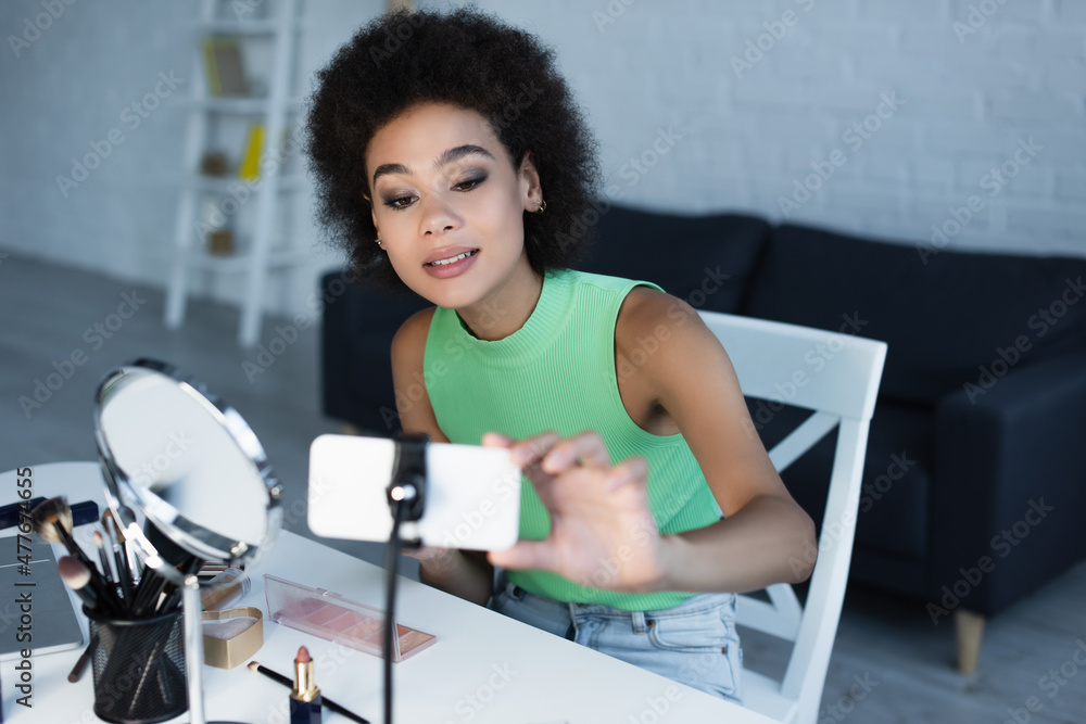 African american blogger using blurred smartphone near decorative cosmetics at home. African american blogger using blurred smartphone near decorative cosmetics at home.