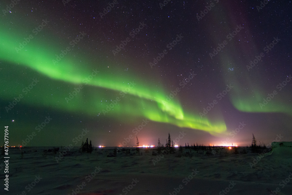The northern lights and aurora borealis fill the sky above distant city lights near Churchill, Manitoba, Canada