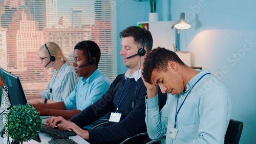 Multiracial call center agent feeling exhausted and broken-down after telephone call with the client. He working with mixed-race team in modern office.