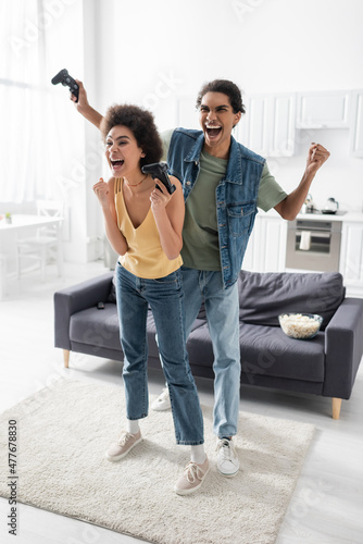 KYIV, UKRAINE - NOVEMBER 9, 2021: Excited african american couple showing yes gesture while holding joysticks at home.
