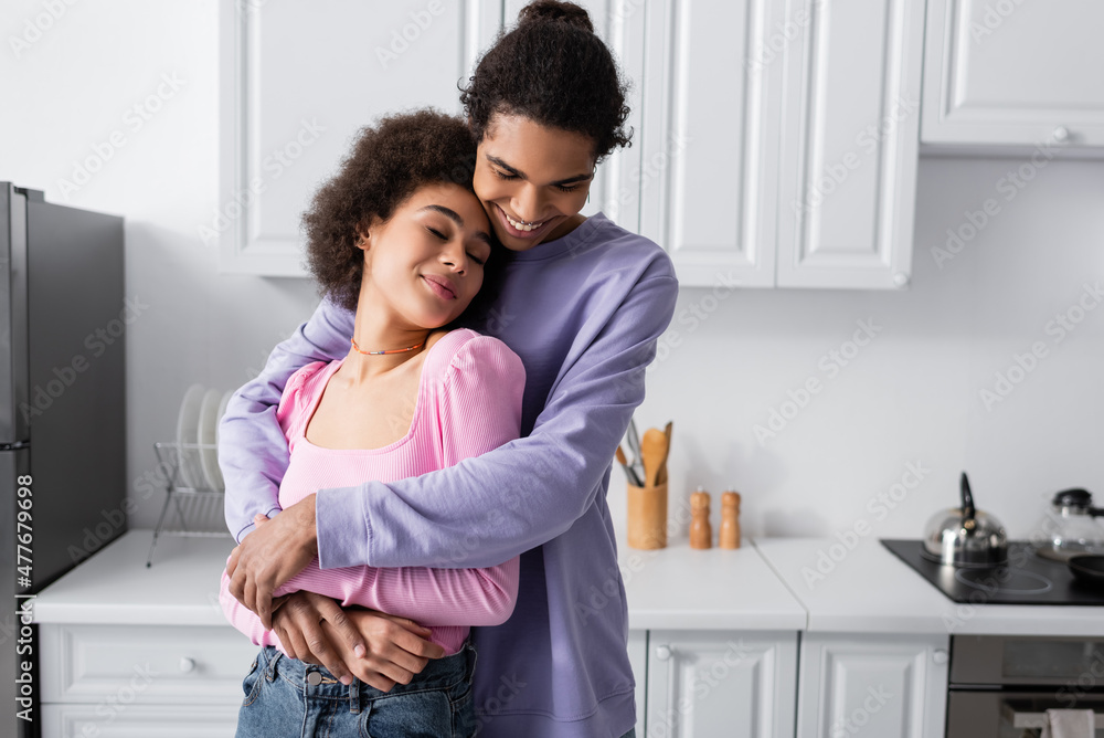Cheerful african american man hugging girlfriend with closed eyes in kitchen.