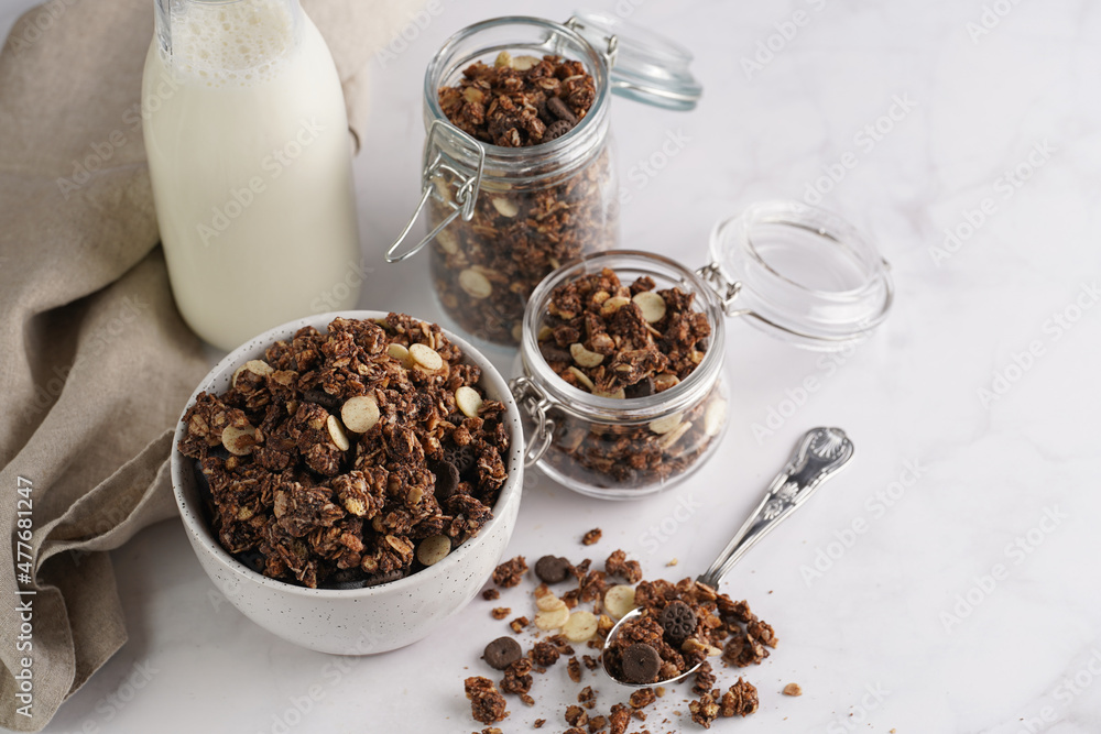 Breakfast muesli cereal with chocolate chips and nuts in several jars and bowl, bottle of milk, white surface Breakfast muesli cereal with chocolate chips and nuts in several jars and bowl, bottle of milk, white surface