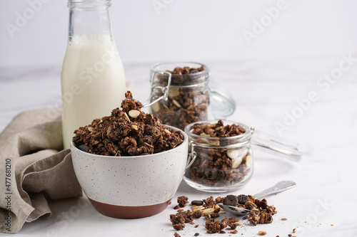 Breakfast muesli cereal with chocolate chips and nuts in several jars and bowl, bottle of milk, white surface