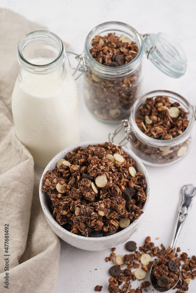 Breakfast muesli cereal with chocolate chips and nuts in several jars and bowl, bottle of milk, white surface Breakfast muesli cereal with chocolate chips and nuts in several jars and bowl, bottle of milk, white surface