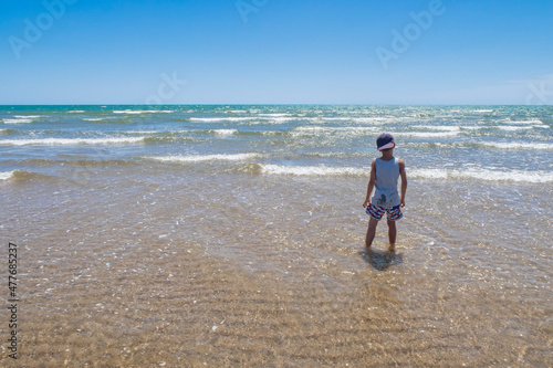 boy with his back to the turquoise water sea on the shore of the beach, small waves wetting his clothes, summer vacation, Isla Canela, Huelva, Spain, Atlantic Ocean