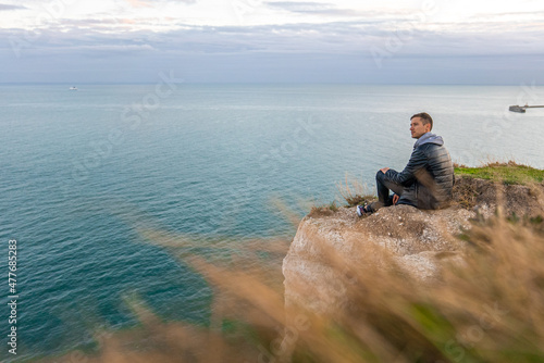 Young man exploring the White Cliffs of Dover in UK. Sitting on top of the cliff watching English canal.