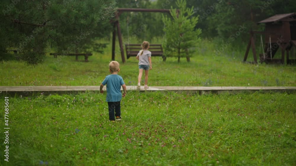 Playful little girl runs to playground with rustic wooden attractions and toddler boy follows sister along meadow backside view slow motion Playful little girl runs to playground with rustic wooden attractions and toddler boy follows sister along meadow backside view slow motion
