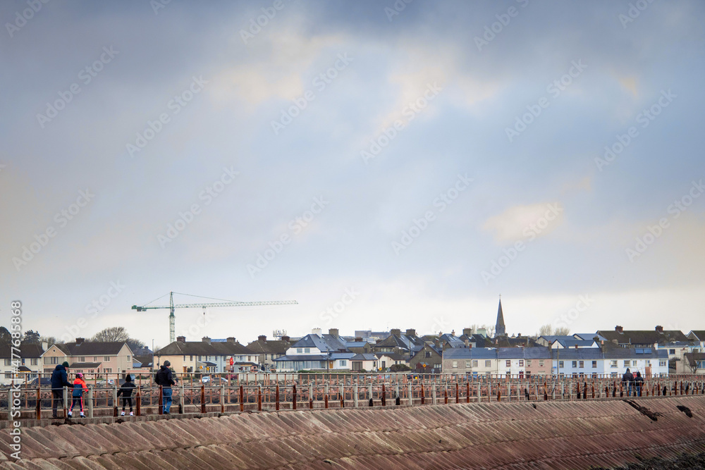 People walking on a Mutton causeway, Galway city in the background. Ireland. Selective focus. Outdoor activity. Cloudy sky. Town buildings in the background. People walking on a Mutton causeway, Galway city in the background. Ireland. Selective focus. Outdoor activity. Cloudy sky. Town buildings in the background.