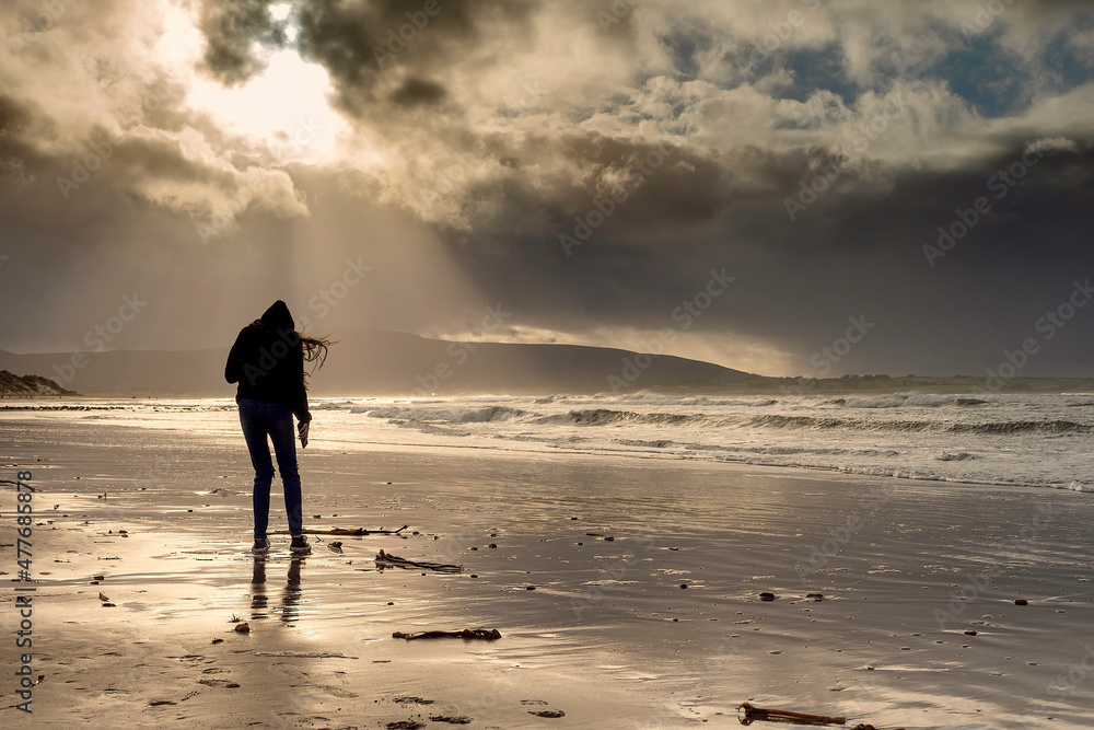 Silhouette of a slim teenager girl walking on a sandy beach. Dramatic cloudy sky in the background, sun flare and burst of light. Outdoor activity. Strandhill beach, county Sligo, Ireland.