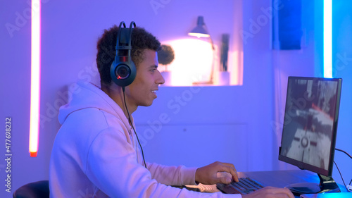 Medium shot of young black gammer in headphones playing in online shooter. The room have warm colored neon light.