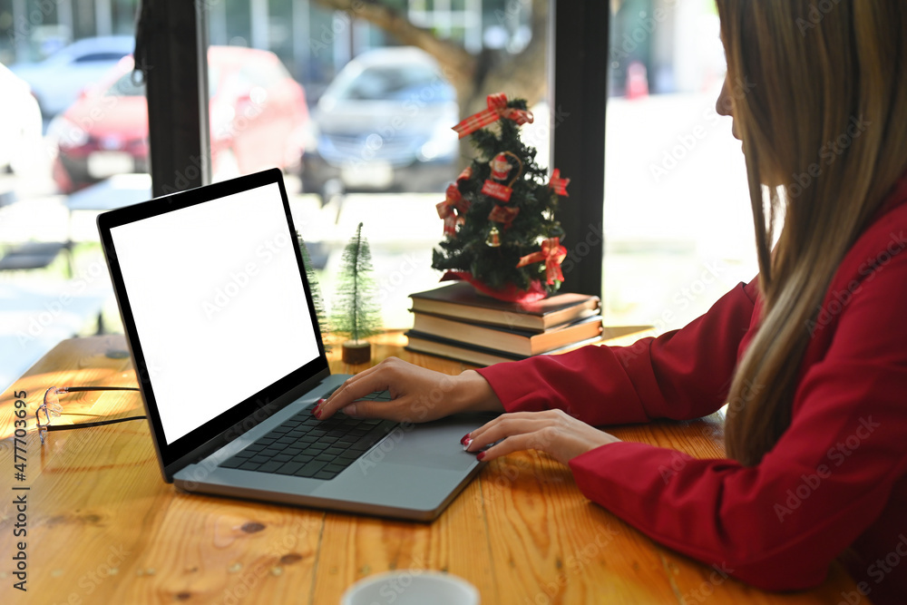 Photo of a young creative woman typing on a computer laptop at the wooden table surrounded by a coffee cup, eyeglasses, stack of books and Christmas tree. Photo of a young creative woman typing on a computer laptop at the wooden table surrounded by a coffee cup, eyeglasses, stack of books and Christmas tree.