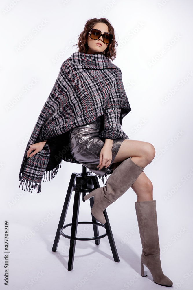 High fashion photo of a beautiful elegant young woman in a pretty gray poncho, metallic silver skirt, suede boots, stylish sunglasses posing over white background. Make up, hairstyle. Slim figure.