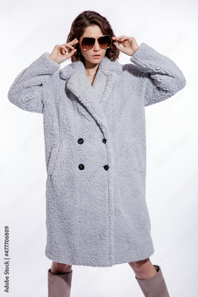 High fashion photo of a beautiful elegant young woman in a pretty pale blue fur coat, suede boots, stylish sunglasses posing over white background. Make up, hairstyle. Slim figure. High fashion photo of a beautiful elegant young woman in a pretty pale blue fur coat, suede boots, stylish sunglasses posing over white background. Make up, hairstyle. Slim figure.