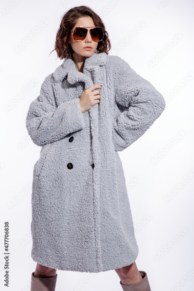 High fashion photo of a beautiful elegant young woman in a pretty pale blue fur coat, suede boots, stylish sunglasses posing over white background. Make up, hairstyle. Slim figure. High fashion photo of a beautiful elegant young woman in a pretty pale blue fur coat, suede boots, stylish sunglasses posing over white background. Make up, hairstyle. Slim figure.