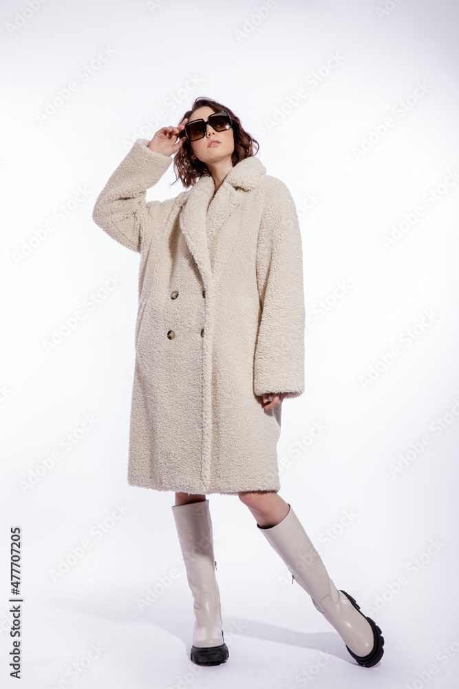 High fashion photo of a beautiful elegant young woman in a pretty cream beige fur coat, boots, stylish sunglasses posing over white background. Make up, hairstyle. Slim figure. High fashion photo of a beautiful elegant young woman in a pretty cream beige fur coat, boots, stylish sunglasses posing over white background. Make up, hairstyle. Slim figure.