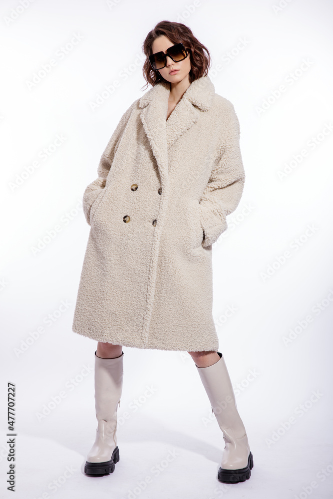High fashion photo of a beautiful elegant young woman in a pretty cream beige fur coat, boots, stylish sunglasses posing over white background. Make up, hairstyle. Slim figure. High fashion photo of a beautiful elegant young woman in a pretty cream beige fur coat, boots, stylish sunglasses posing over white background. Make up, hairstyle. Slim figure.