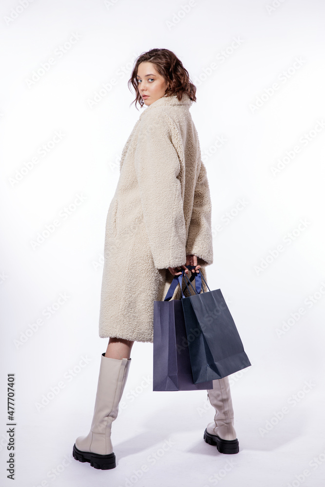 High fashion photo of a beautiful elegant young woman in a pretty cream beige fur coat, boots, two blue shopping bags posing over white background. Make up, hairstyle. Slim figure. High fashion photo of a beautiful elegant young woman in a pretty cream beige fur coat, boots, two blue shopping bags posing over white background. Make up, hairstyle. Slim figure.
