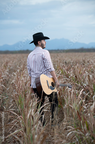 JOVEN MEXICANO CANTANDO CON SU GUITARRA USA SOMBRERO Y TIENE BARBA