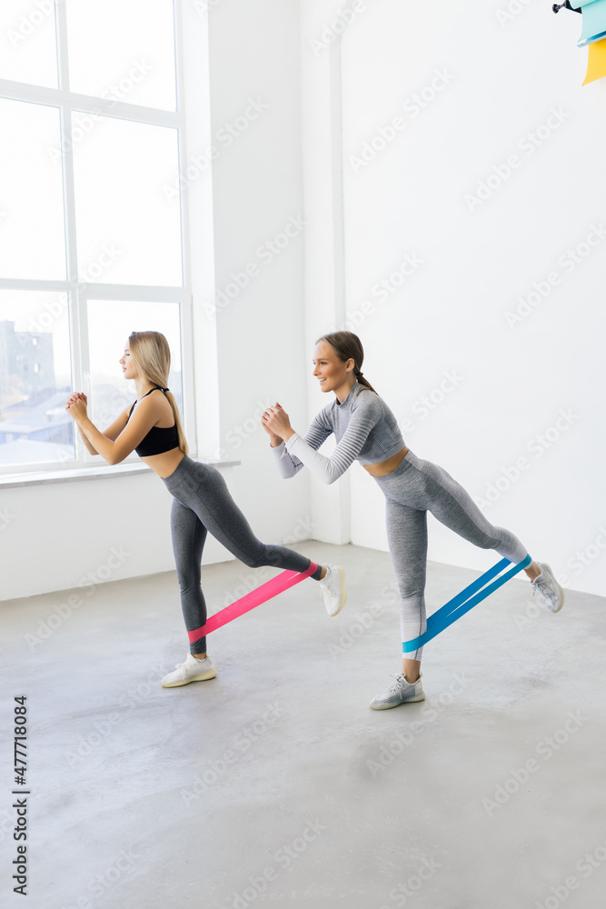 Obraz premium Two smiling athletic women squatting synchronously with outfit in fitness studio. Women are enjoying leg and buttocks work out with resistance band. Training together with friend in gym concept